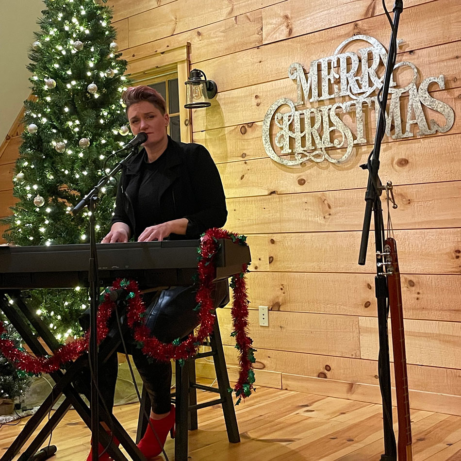 Chelsea Berry at the piano, merry christmas sign in the corner and a Christmas tree behind her.